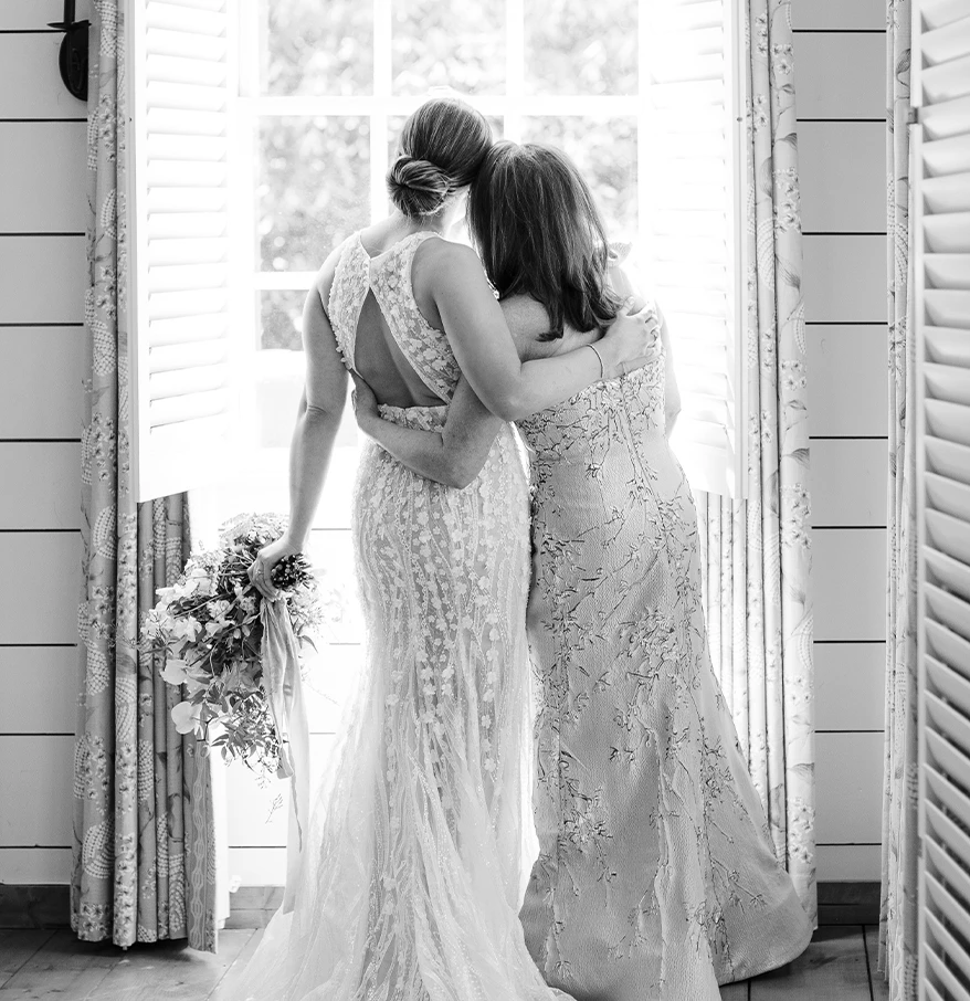two women in dresses hugging in front of a window