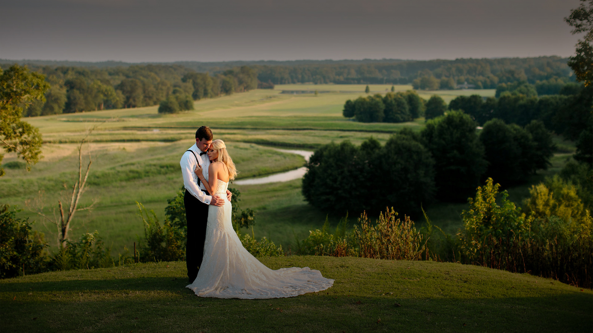a man and woman in wedding attire hugging on a hill with trees and a river