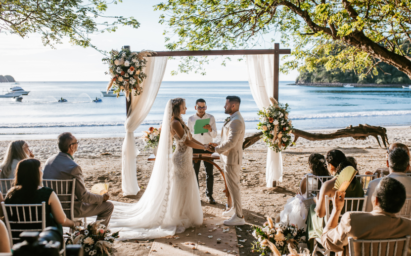 a couple in white wedding dress under a wooden arch with people sitting in chairs and trees