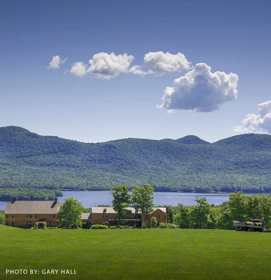 a house in a grassy field with trees and a lake in the background