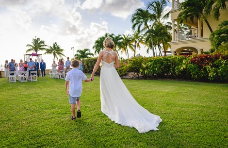 a woman in a white dress walking down the grass with a boy holding hands