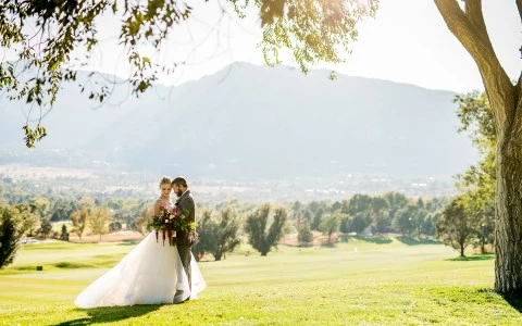 a man and woman in wedding attire standing in a field with trees and mountains in the background