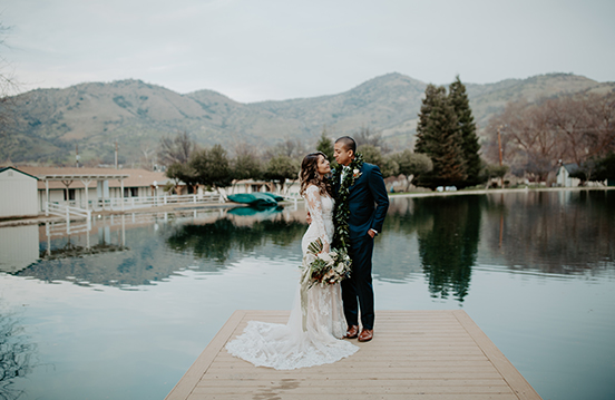 a man and woman kissing on a dock near a lake